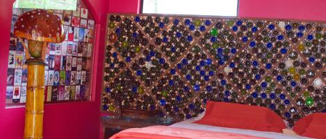 A room with a wall mosaic of glass bottles, a bed with red bedding, and a bamboo lamp.
