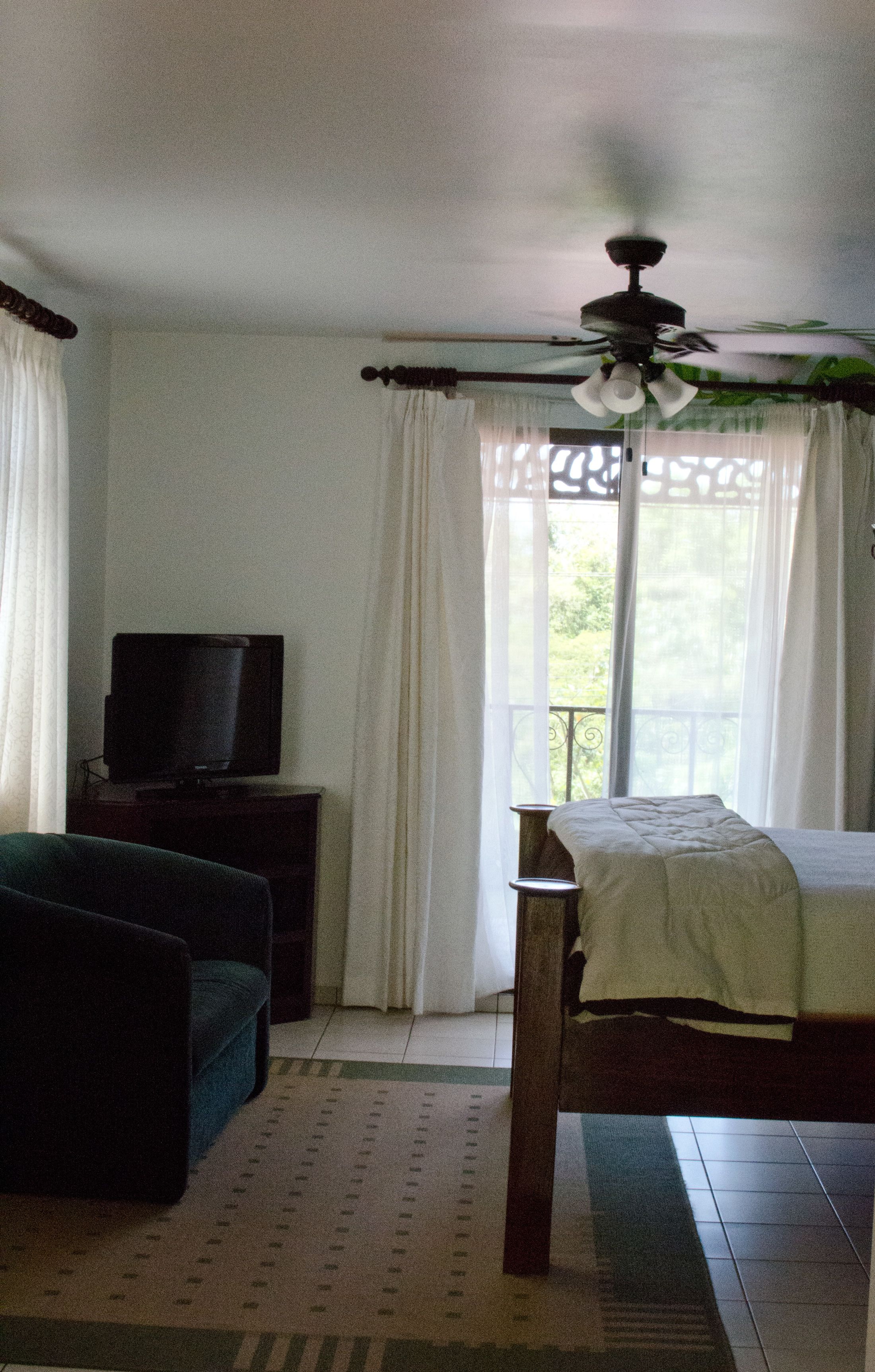 A bedroom with a bed, a television on a stand, a ceiling fan, and a window with curtains.
