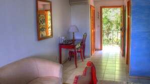 Quarto com sofá, escrivaninha com abajur e cadeira. Porta dá acesso a área externa com vista para vegetação.
