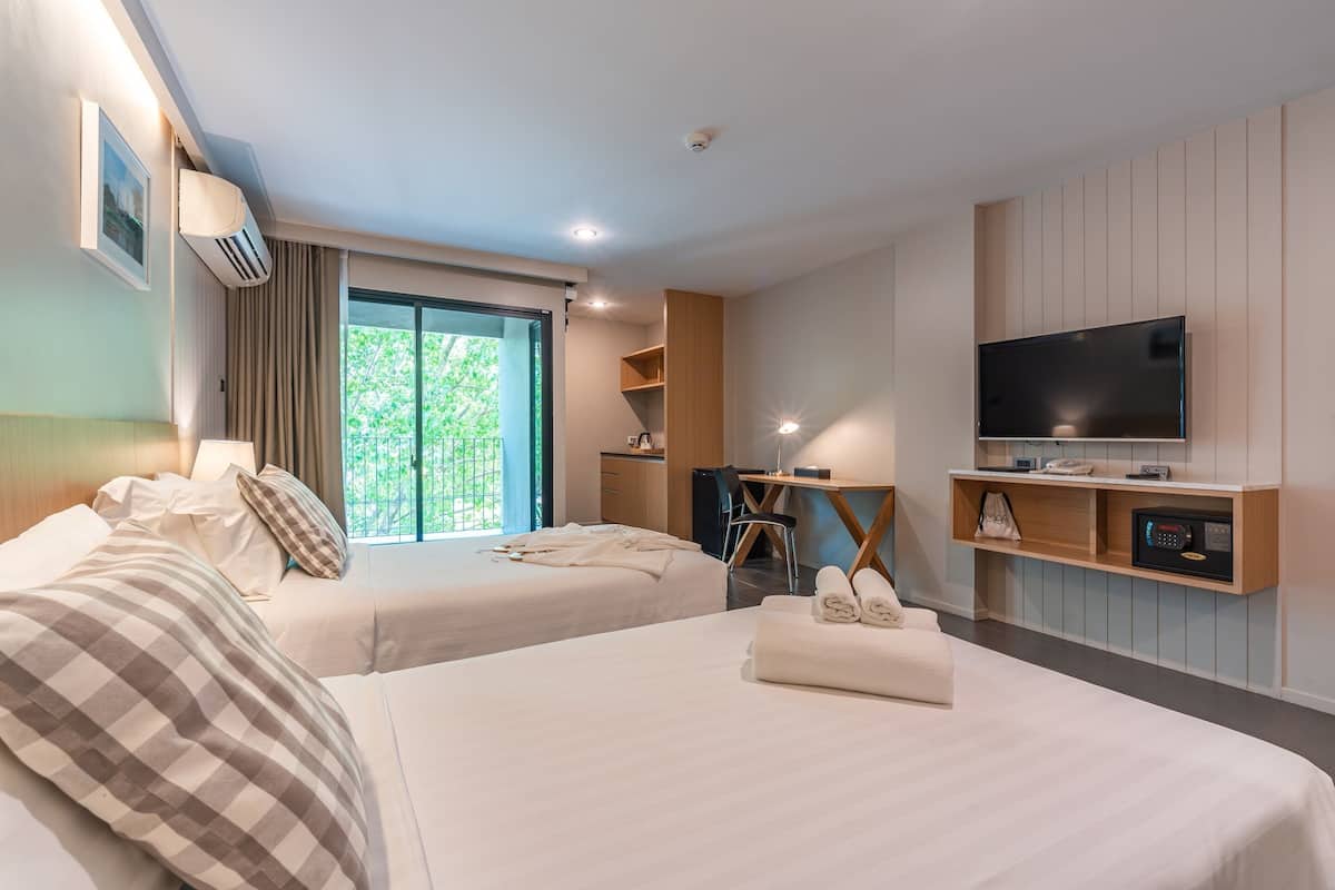 A hotel room with two beds, a TV, a desk, and a window with a view of greenery.