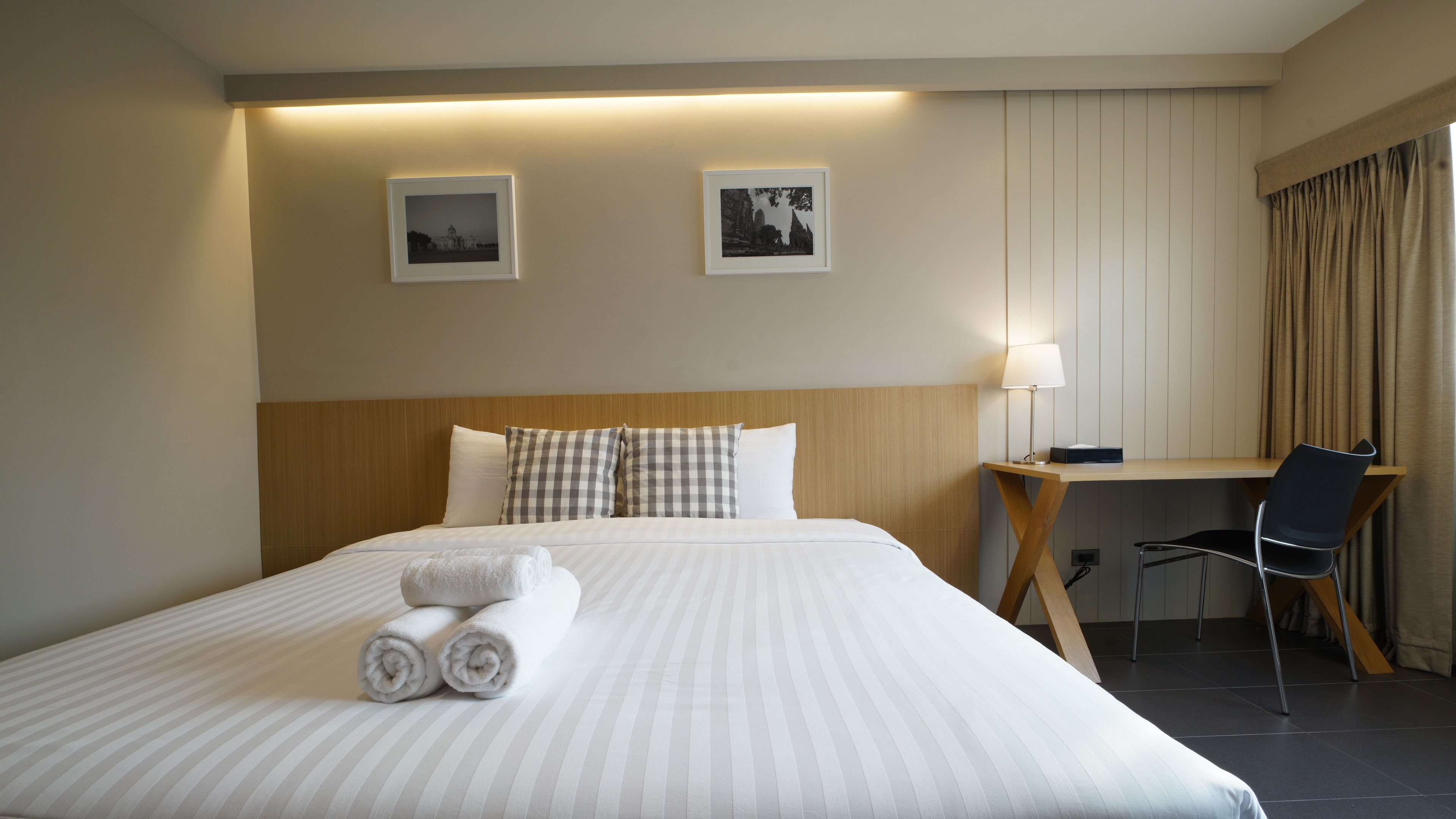 A hotel room with a neatly made bed, two framed pictures on the wall, a desk with a chair, and a lamp.