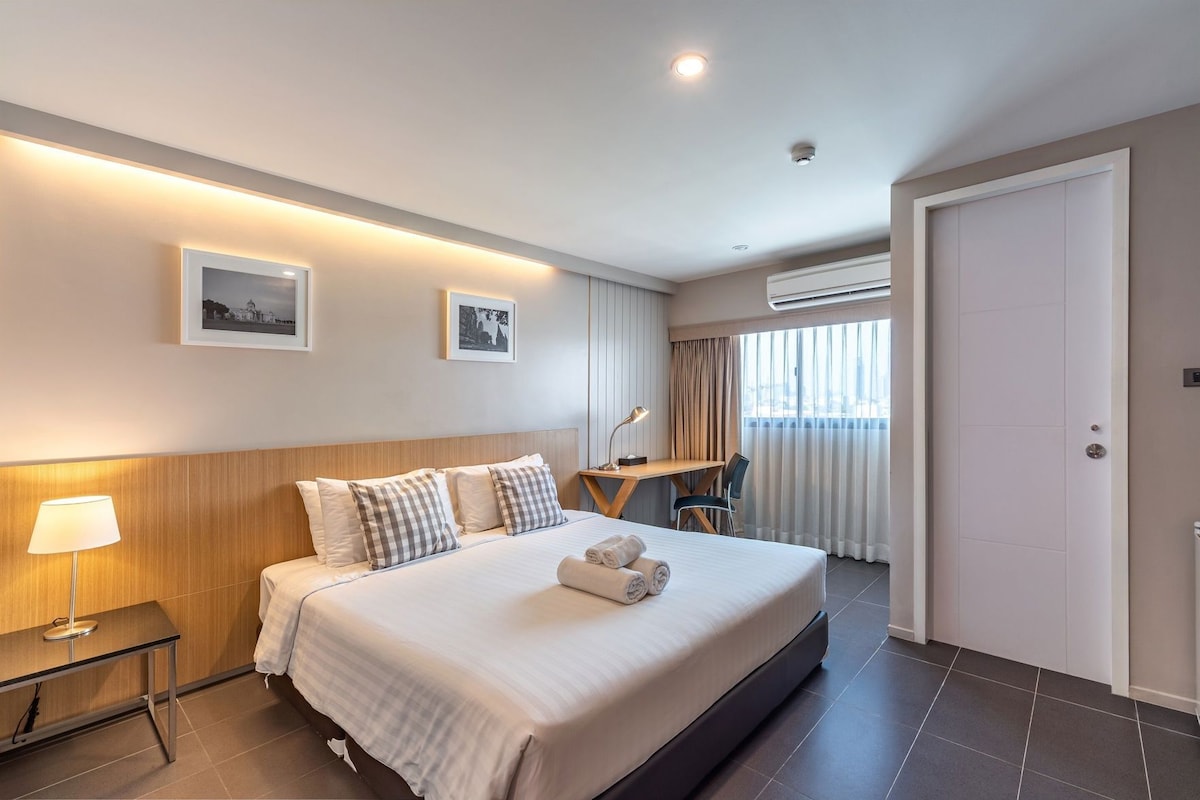 A hotel room with a bed, bedside table, lamp, desk, chair, and an air conditioning unit.