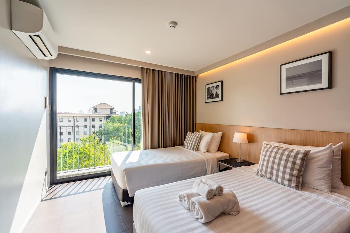 A hotel room with two beds, a large window with curtains, a wall-mounted air conditioner, and a framed picture.