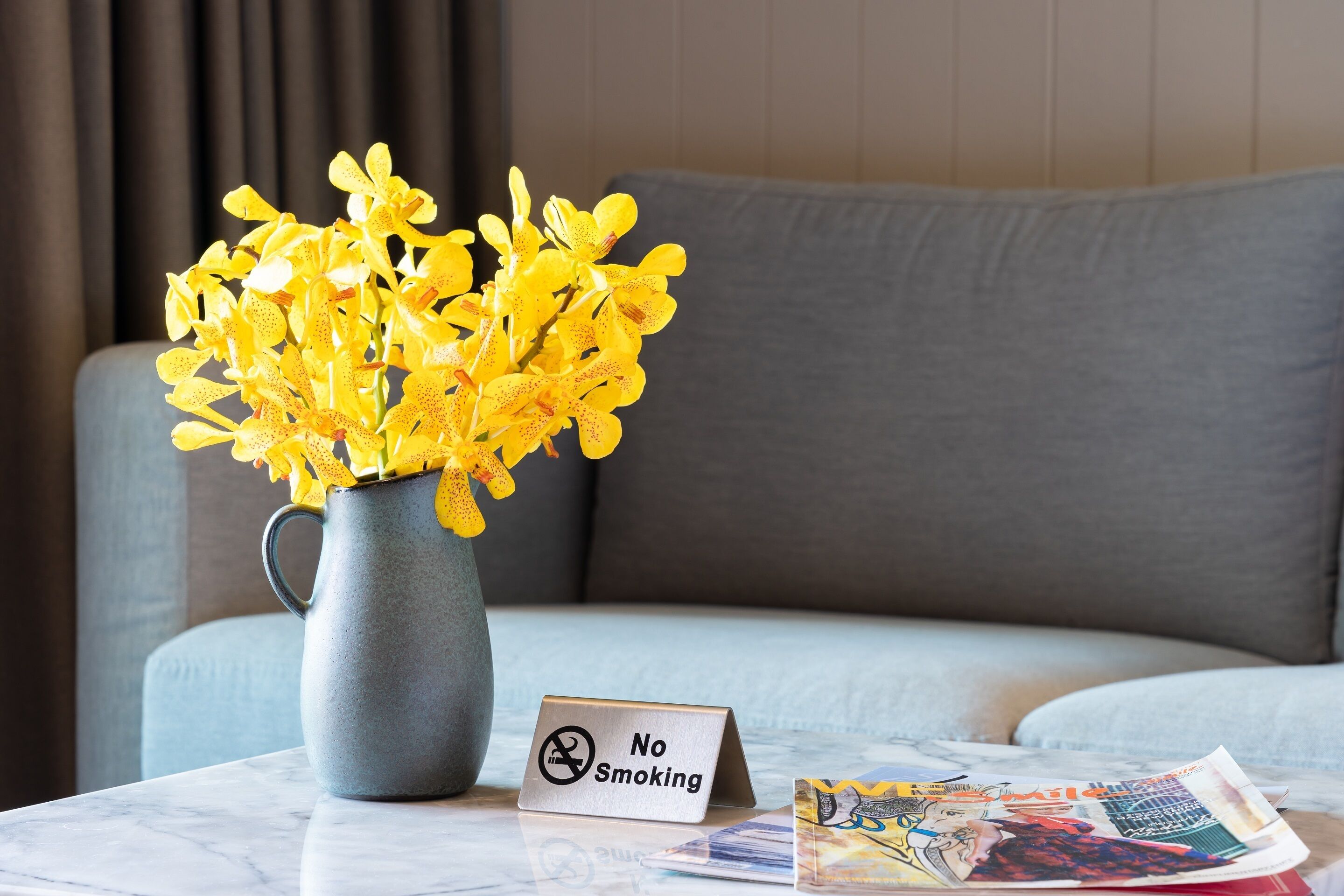 Eine graue Vase mit gelben Blumen auf einem Marmortisch, ein 'No Smoking'-Schild und eine Zeitschrift.