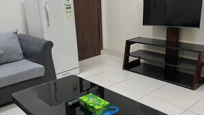 A modern living room with a glass-top coffee table, a grey sofa, a white refrigerator, a wooden door with notices, and a mounted TV. - Binayat Hama Serviced Apartments (Al Jubail)