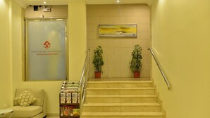A hotel reception area with a staircase, a sofa, a glass door with a logo, and two potted plants. - Binayat Hama Serviced Apartments (Al Jubail)