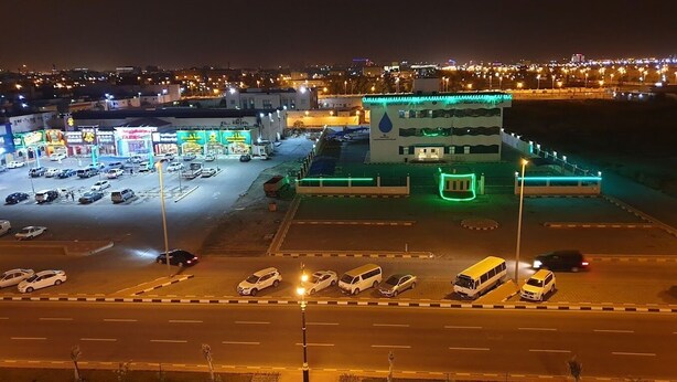 A parking lot with multiple cars and a building with illuminated signage. - Binayat Hama Serviced Apartments (Al Jubail)