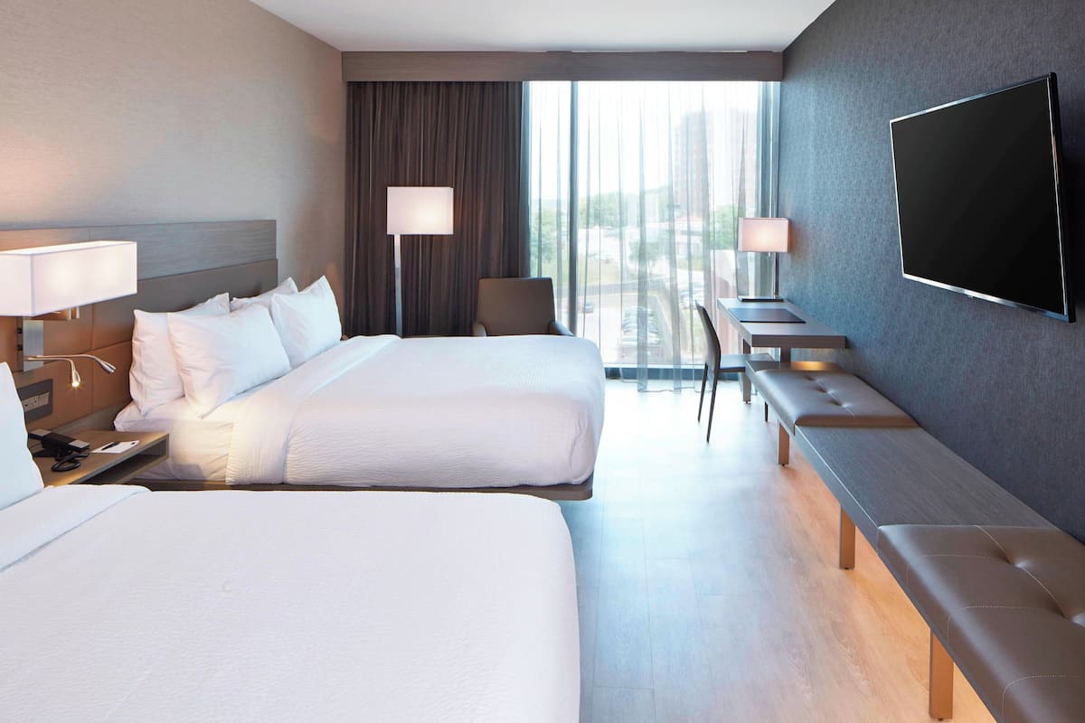 Habitación de hotel con dos camas, televisión, escritorio con silla y un ventanal con cortinas.