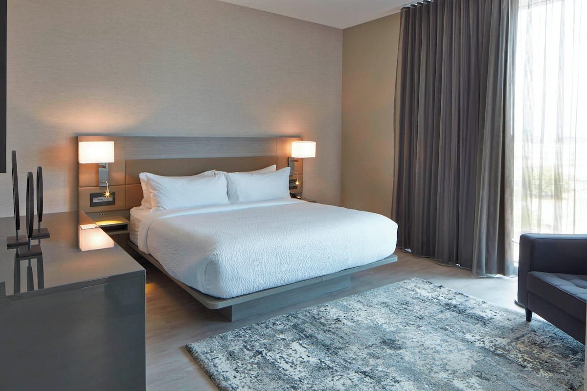 Habitación de hotel moderna con una cama grande, dos lámparas de noche, un sofá de color oscuro y un ventanal con cortinas.