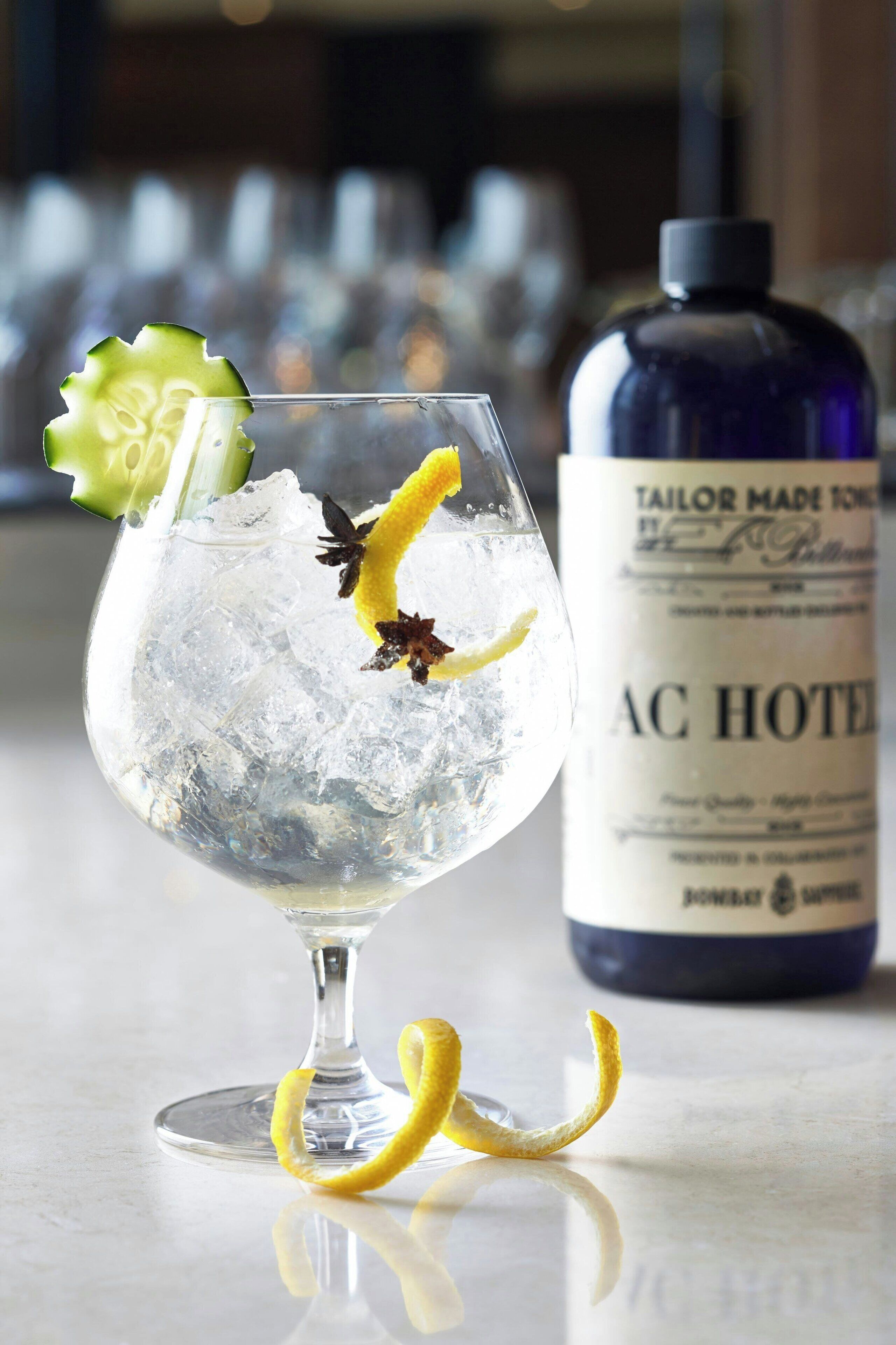A cocktail glass with ice, lemon peel, and a cucumber garnish on a bar counter. A bottle labeled "Tailor Made Tonics" is visible in the background.