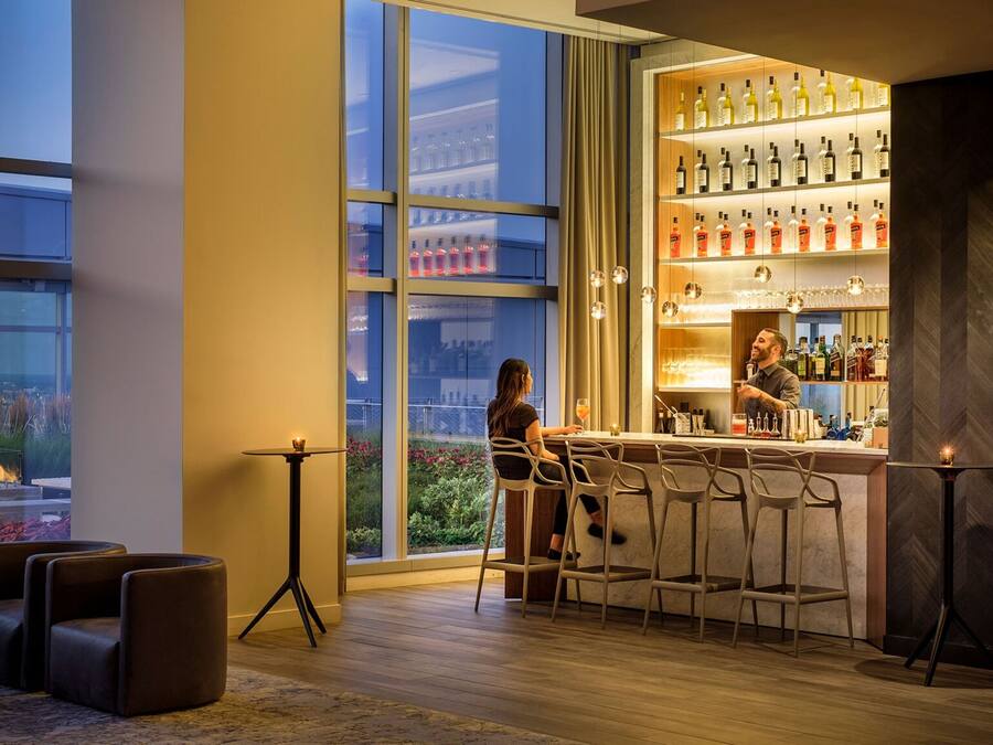 A modern bar area with a bartender, a woman seated at a table, and a large window offering a city view.