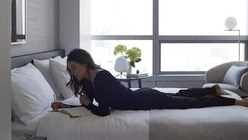 A modern hotel room with a large bed, a bedside table, a small table with a plant, and a view of the city through large windows.
