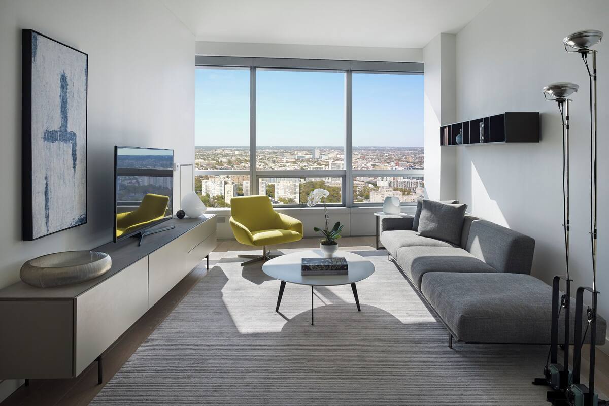 Un salon moderne avec vue sur la ville, comprenant un canapé, un fauteuil jaune, une table basse et un téléviseur.