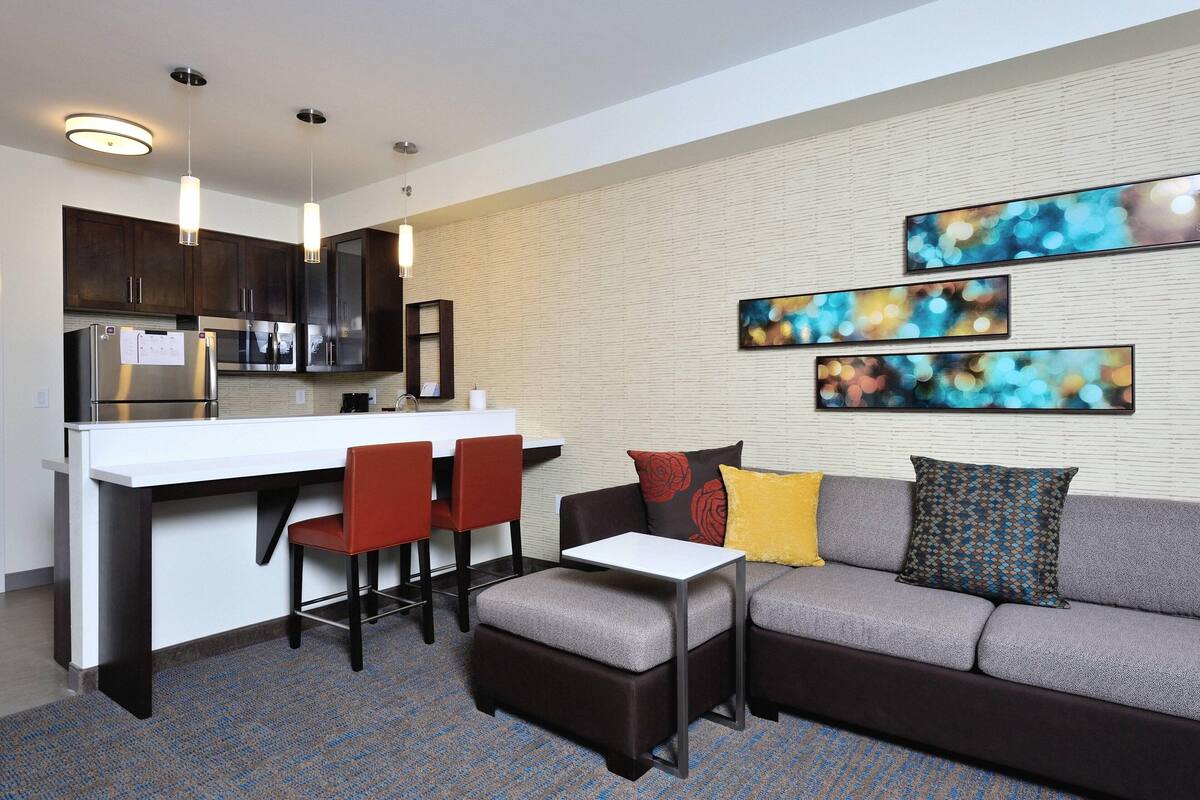 A modern kitchen with dark cabinets, a white countertop, and a stainless steel refrigerator. There is a dining area with a gray sofa, red chairs, and a small white table. The room has a patterned carpet and abstract art on the wall.