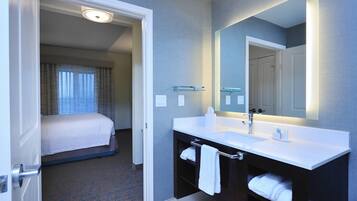 Habitación de hotel con cama, baño con lavamanos y espejo blancos, y pasillo que conduce a otra habitación.