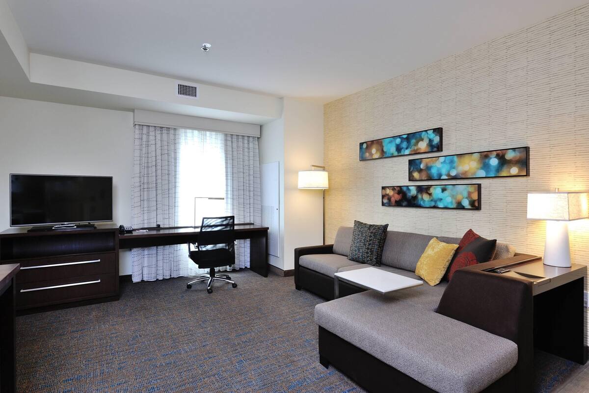 A hotel room with a flat-screen TV, a desk with a chair, a sofa with cushions, and two wall-mounted lamps.
