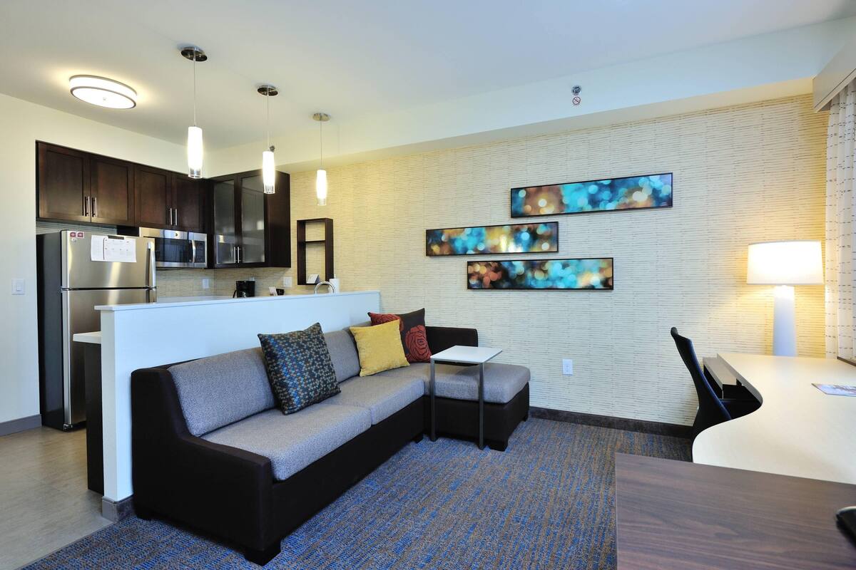 A modern kitchen with dark cabinets, stainless steel appliances, and pendant lighting. There is a seating area with a sofa and a small table, and artwork on the wall.