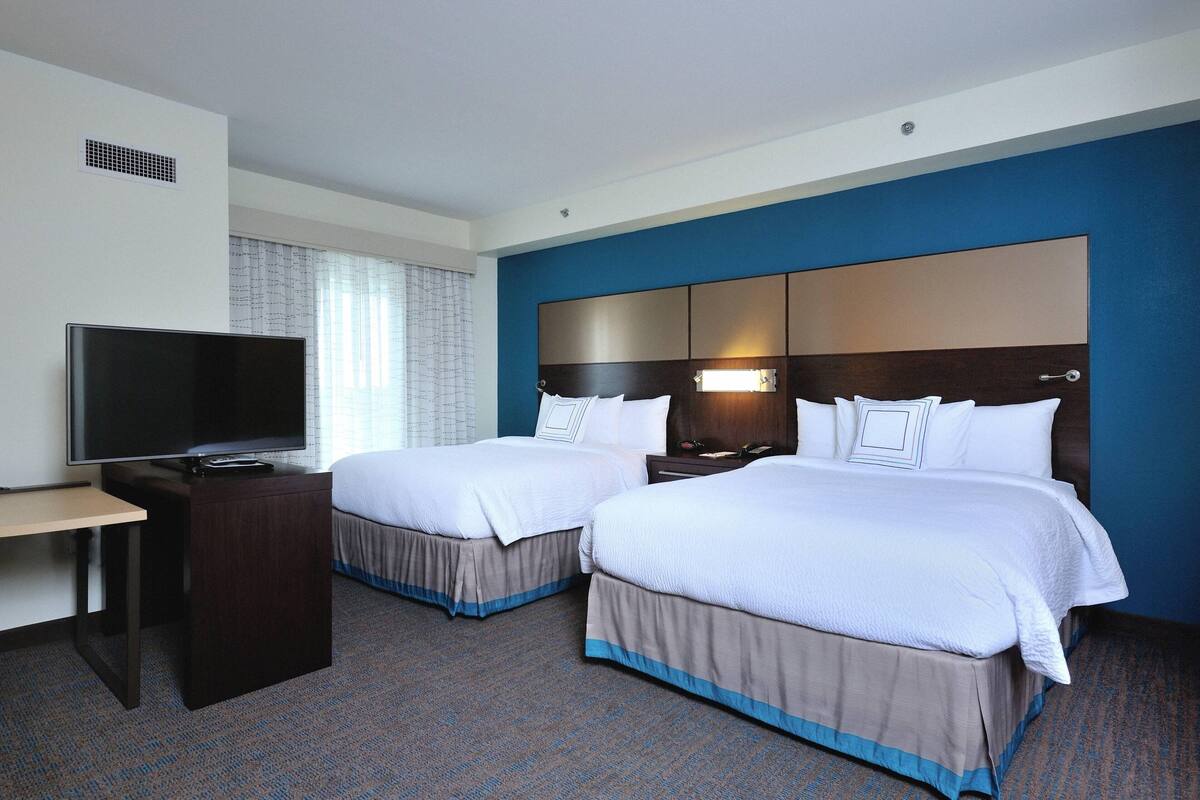 A hotel room with two beds, a TV, a desk, and a window with curtains.