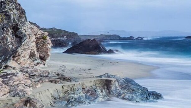 A coastal scene with a rocky cliff, sandy beach, and ocean waves. - The Beach Day's Bar & B&B (Inishbofin)