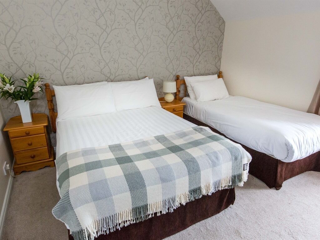 Un dormitorio con dos camas individuales, una mesita de noche con lámpara, un jarrón con flores y un papel tapiz estampado.