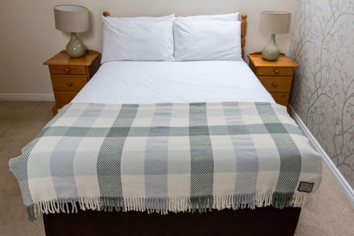 A bed with white linens, flanked by two nightstands with lamps, a plaid blanket on the bed, and a patterned wallpaper on the wall.