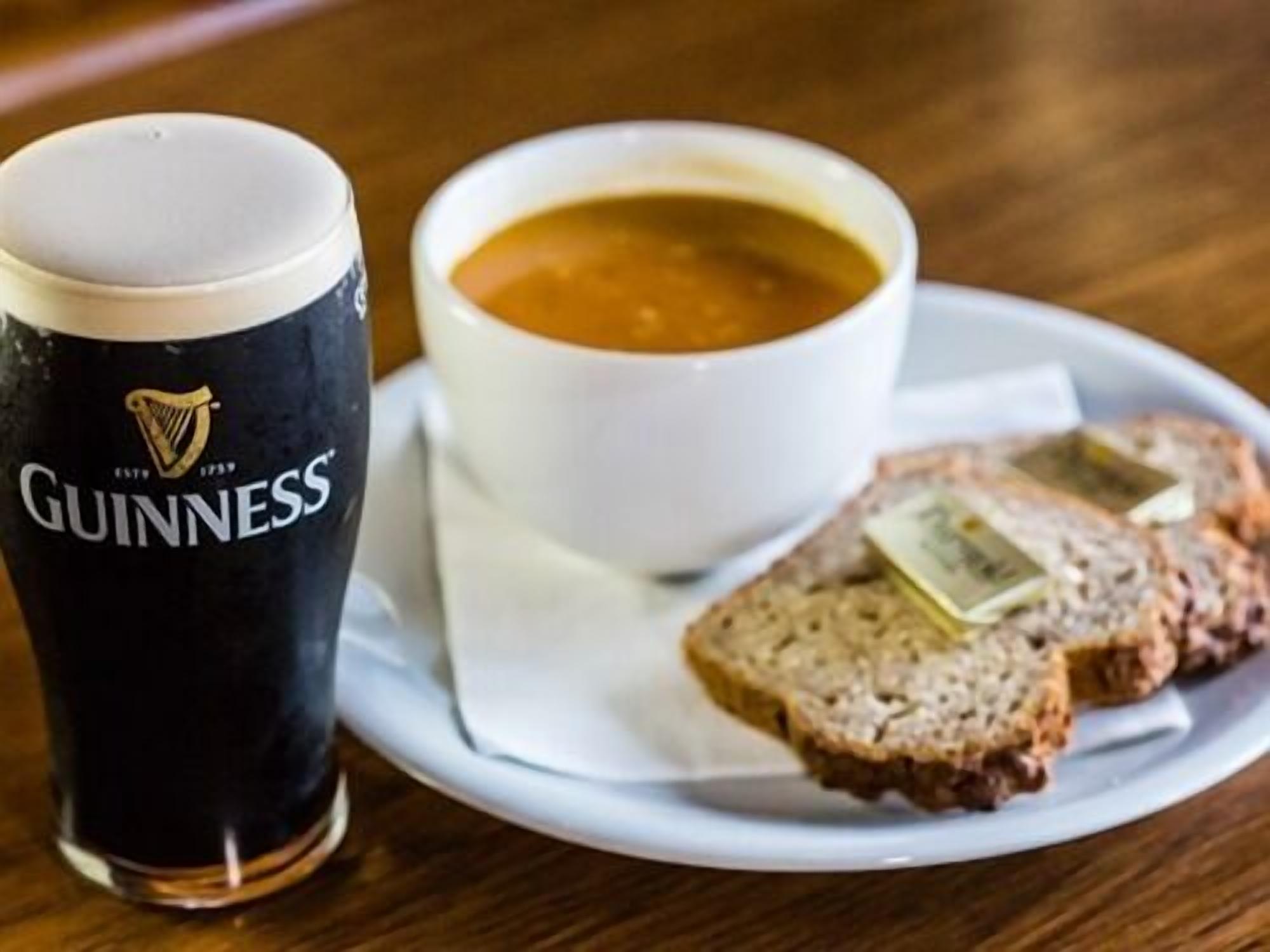 Un verre de bière Guinness, un bol de soupe et des tranches de pain sur une assiette blanche.