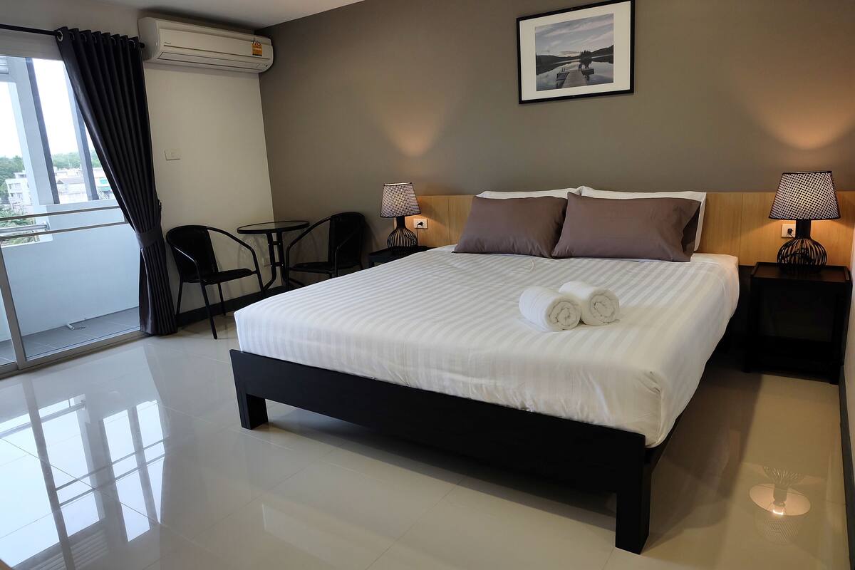 A hotel room with a large bed, two bedside lamps, a small table, a chair, and a view of the outdoors through a window.