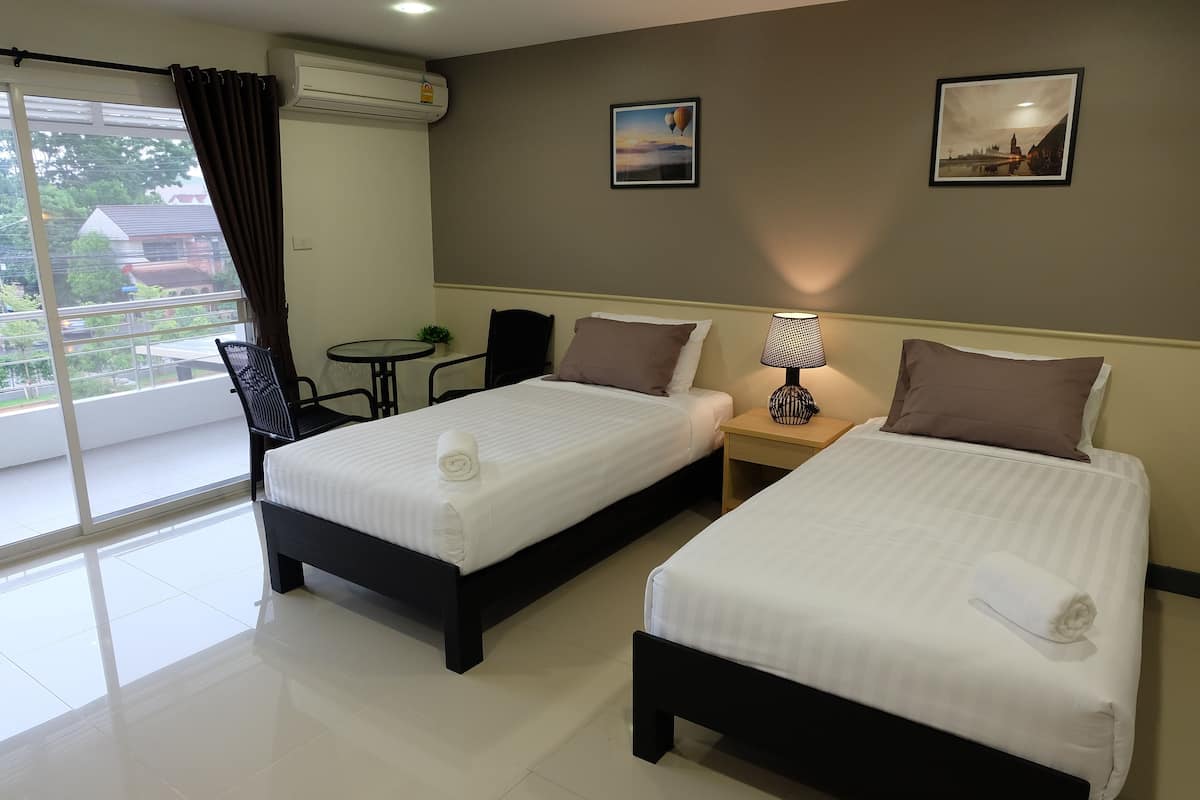 A hotel room with two beds, a small table, a chair, a window with curtains, and a wall-mounted air conditioner.