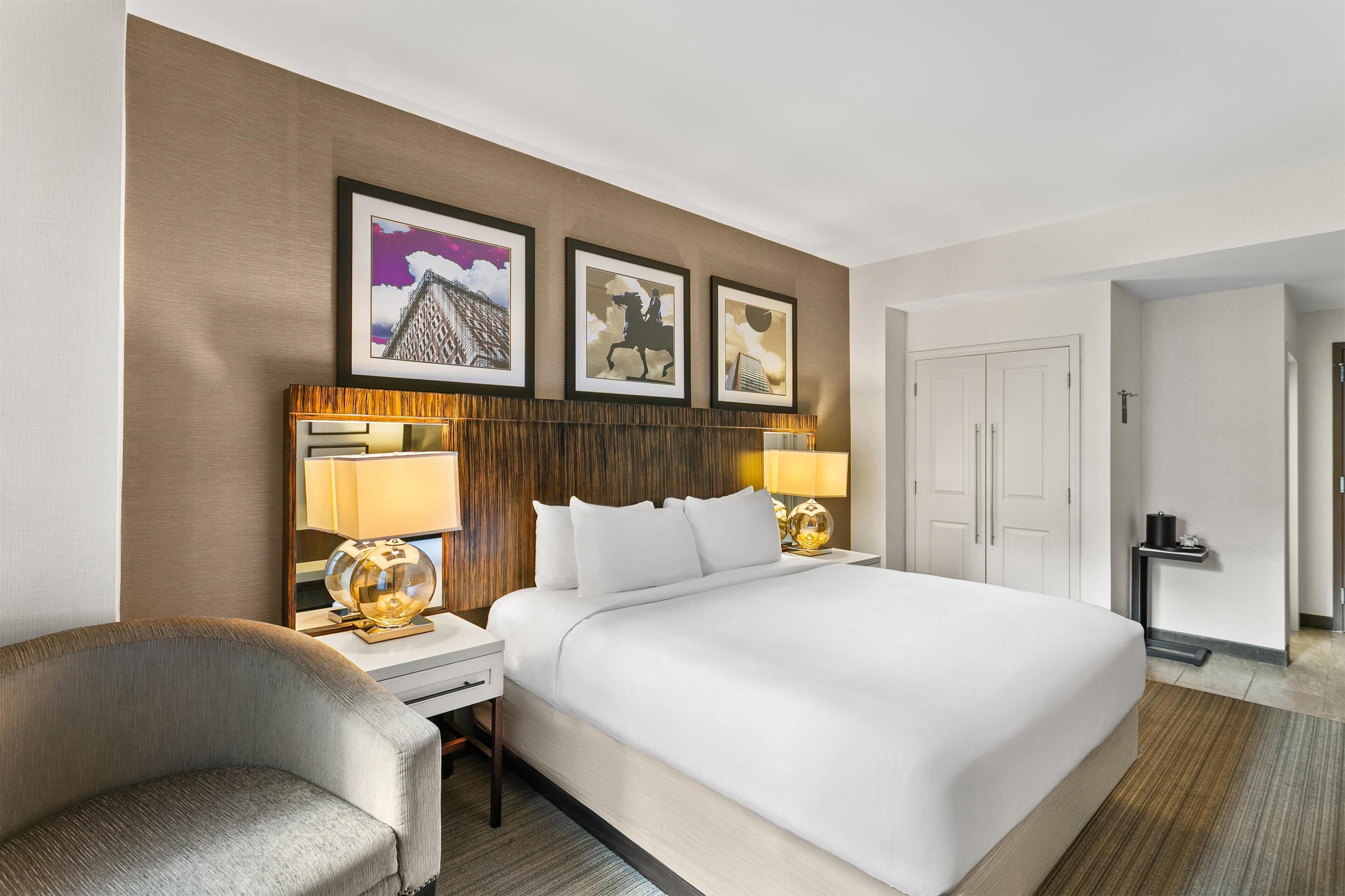 A hotel room with a large bed, two bedside tables with lamps, a chair, and framed artwork on the wall.
