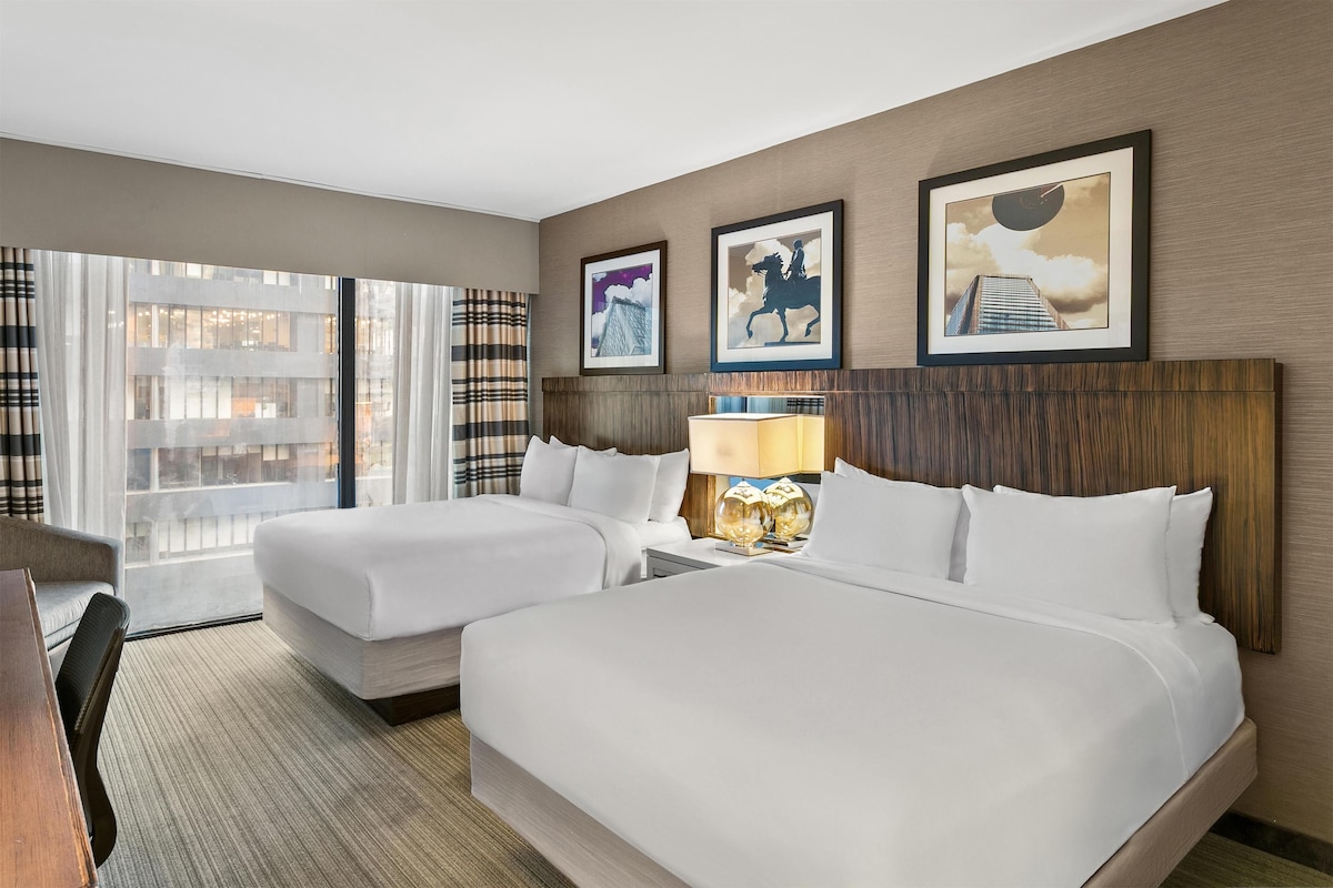 A hotel room with two beds, a desk, a chair, a lamp, and framed artwork on the wall.