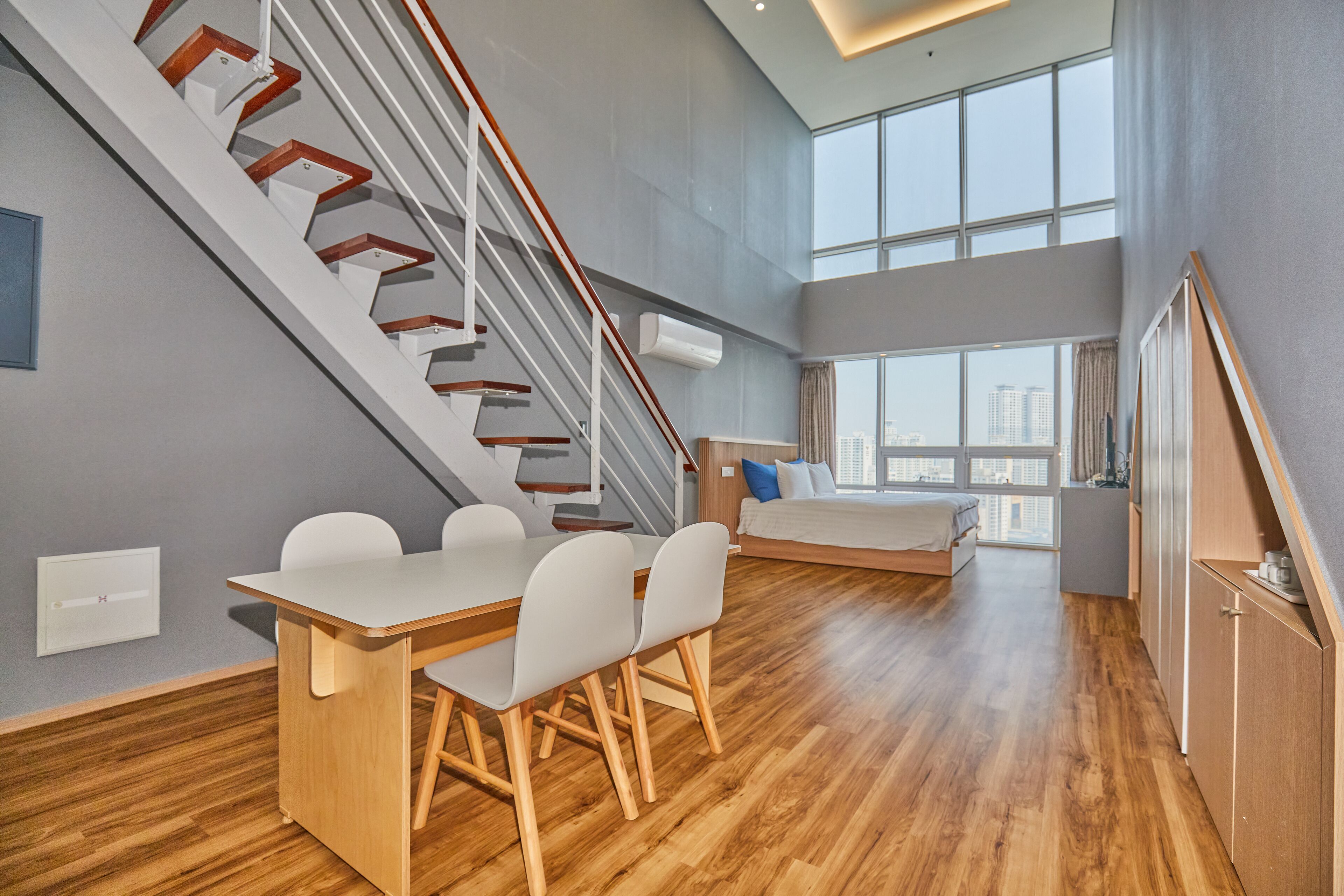 A modern apartment with a wooden dining table, white chairs, a staircase, a bed with a city view, and a large window.