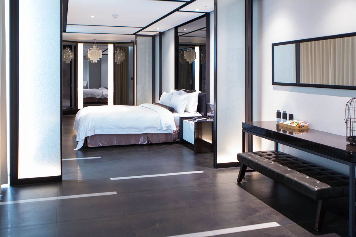 A modern hotel room with a large bed, a bench, a desk, and a mirror.