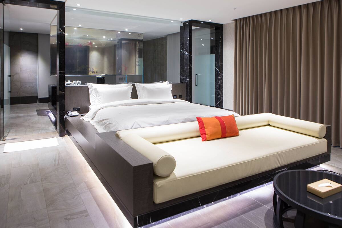 A modern hotel room with a large bed, a bedside table, a mirror, and a view of the cityscape.