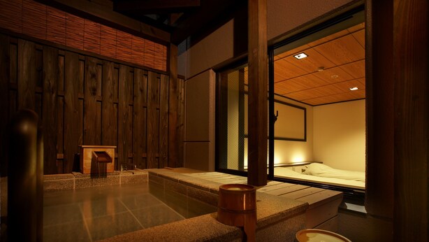 Una habitación de estilo japonés tradicional, con suelo de tatami, un banco de madera, una pequeña mesa de madera y una puerta corredera que da a la zona de la cama.