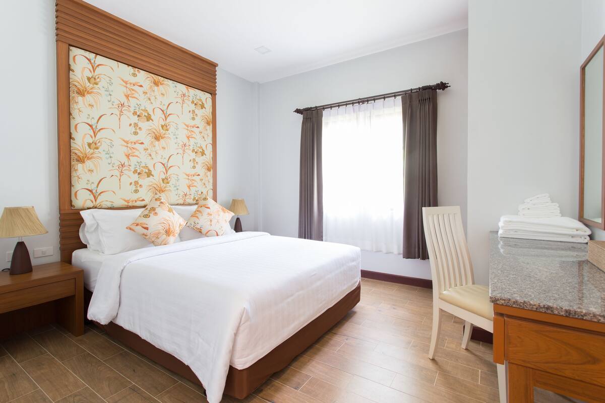 A hotel room with a large bed, a wooden headboard, a nightstand with a lamp, a chair, a desk with a mirror, and a window with curtains.