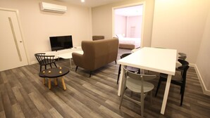 Una habitación de hotel moderna con piso de madera, una mesa de comedor blanca, un sillón marrón, un televisor y una cama con vista al exterior.