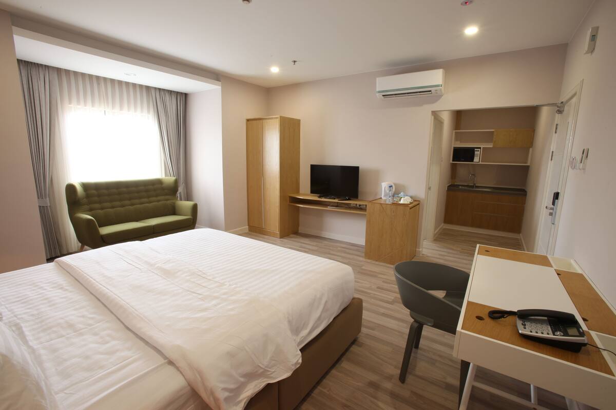Quarto de hotel com cama, sofá verde, escrivaninha com telefone, televisão e cozinha compacta.