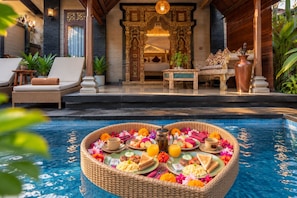 A poolside breakfast setup with a wicker tray filled with food and drinks, a bed with ornate wooden headboard, and a lounge area with wicker furniture. - Ashanti Villa Ubud (Ubud)