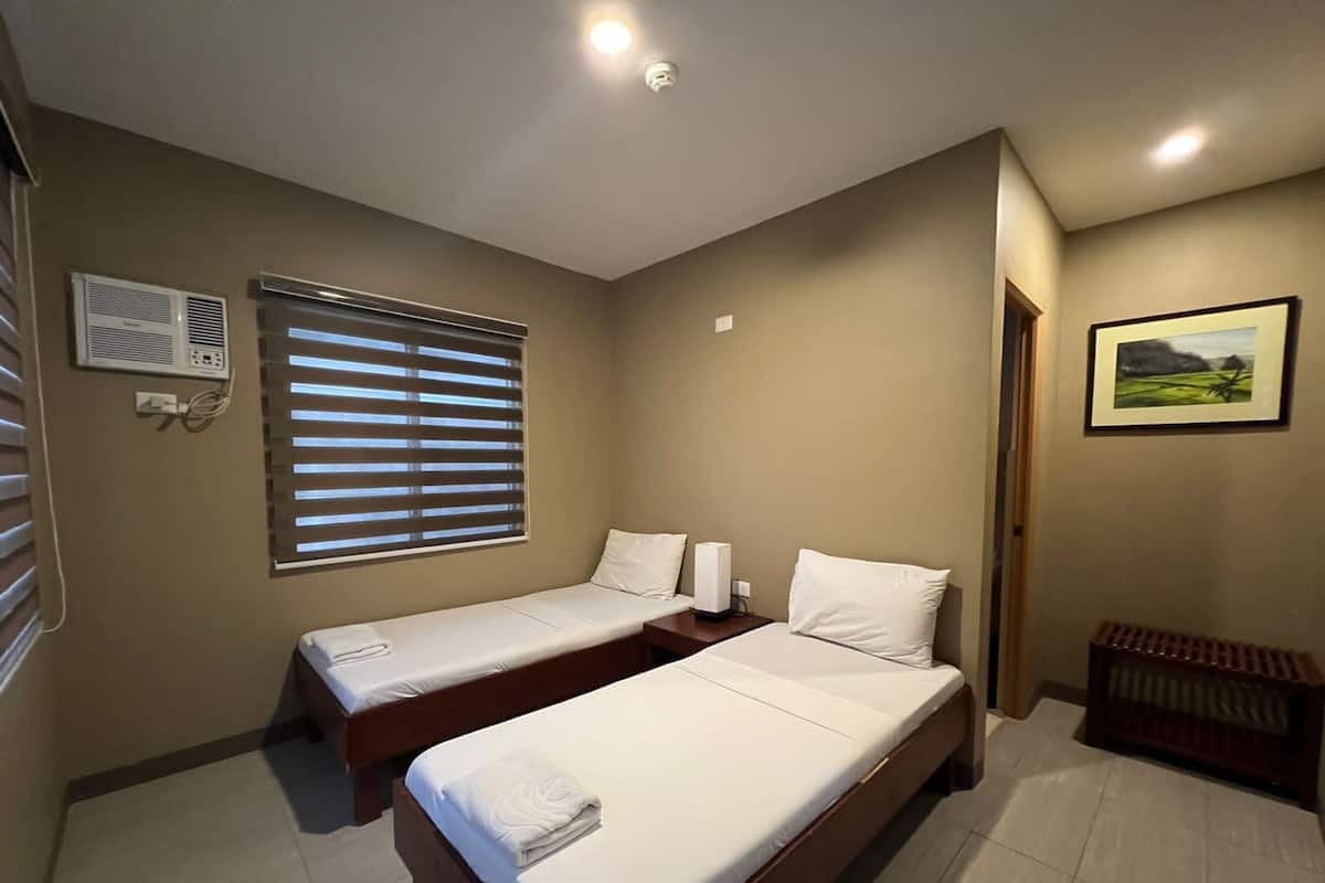 A hotel room with two single beds, a window with blinds, a wall-mounted air conditioner, and a framed picture on the wall.