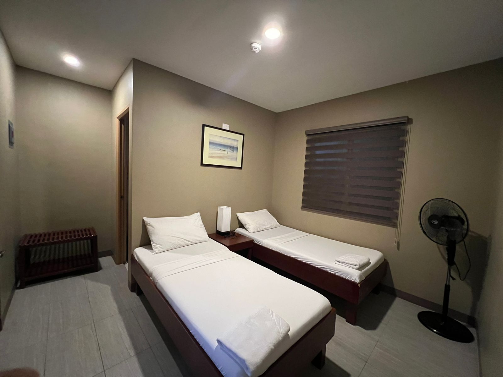 A hotel room with two single beds, a standing fan, a radiator, a framed picture on the wall, and a window with blinds.