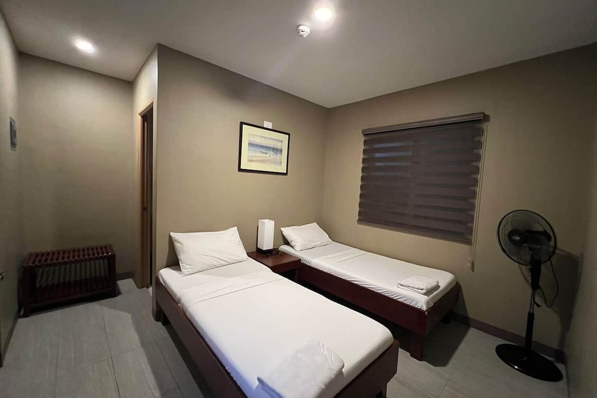 A hotel room with two single beds, a standing fan, a radiator, a framed picture on the wall, and a window with blinds.