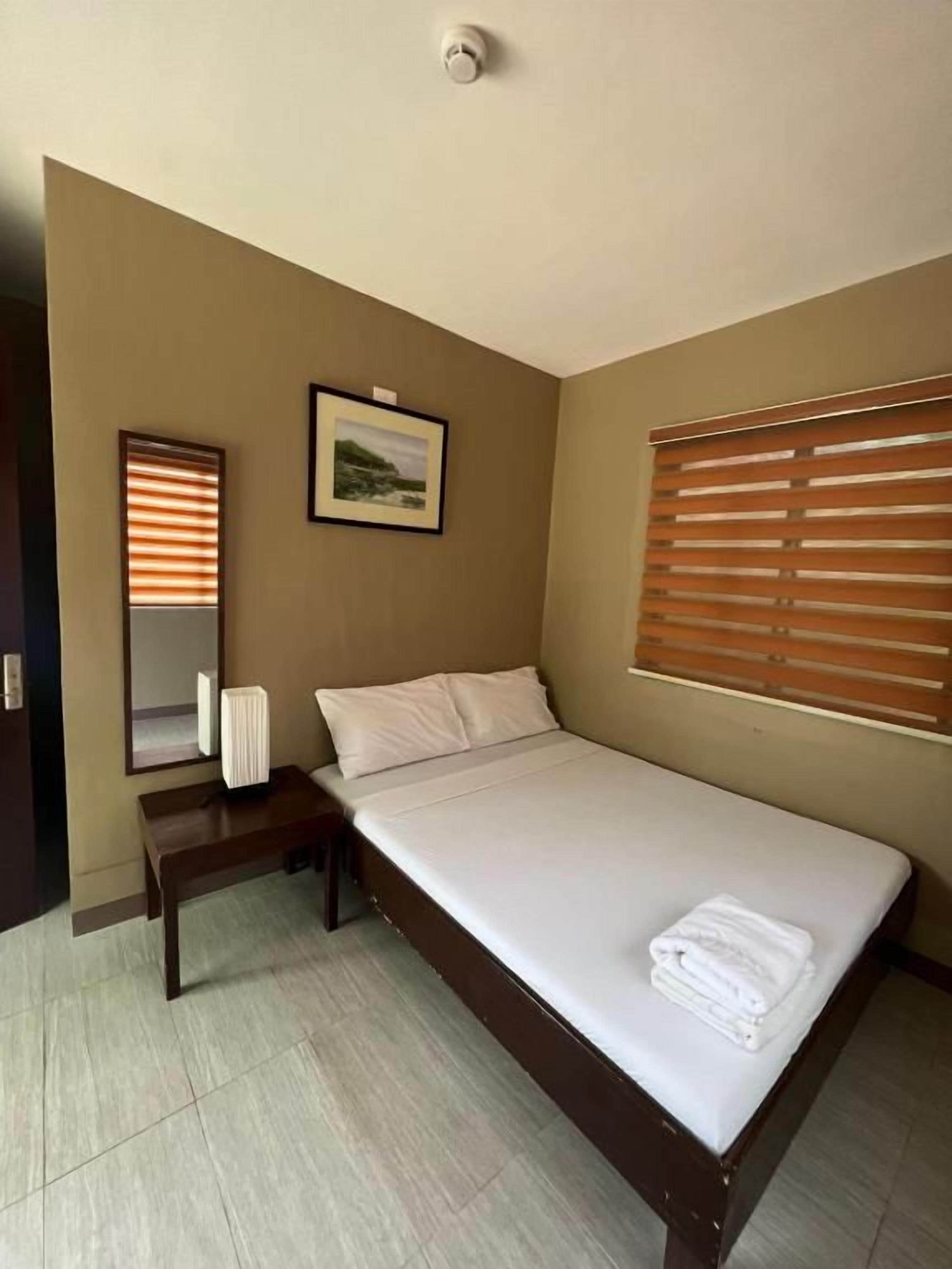 A single bed with a wooden frame and white bedding, a bedside table with a lamp, a wall-mounted mirror, and a window with blinds.
