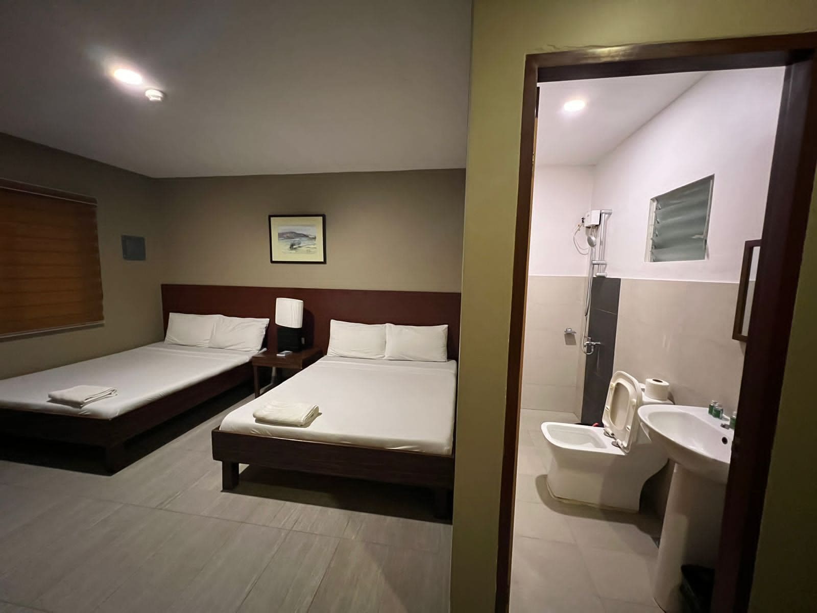 A hotel room with two single beds, a bedside table with a lamp, a bathroom with a toilet, sink, and shower.
