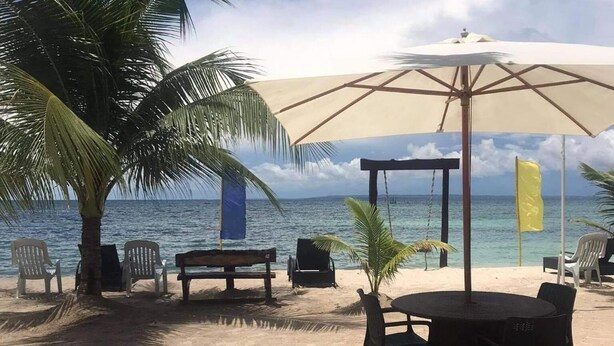 A beachside seating area with a table and chairs, a large umbrella, and a palm tree. - Anaya Beach Resort (Bantayan)