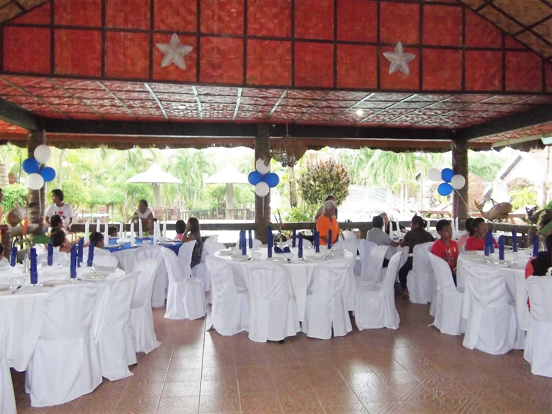 A covered outdoor dining area with white tablecloths and blue and white balloons.