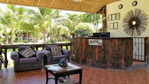Zona de recepción con un escritorio de madera, dos sillones acolchados, una mesa de centro y vistas a palmeras.