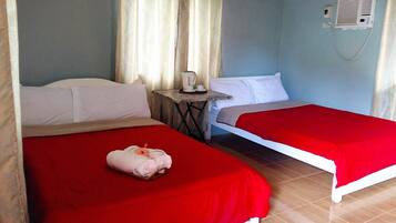 A hotel room with two single beds, each with a red bedspread, a small table with a kettle, and a wall-mounted air conditioning unit.