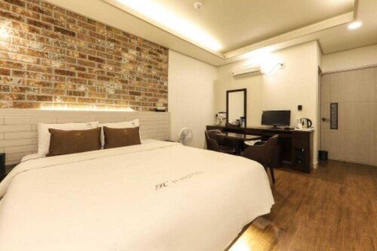 Una habitación de hotel con pared de ladrillo decorativa, una cama grande con ropa de cama blanca, un escritorio con silla y un televisor de pantalla plana.