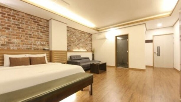 A modern hotel room with a brick accent wall, a large bed, a bench, a small table, and a door. - H Hotel Ulsan (Ulsan)