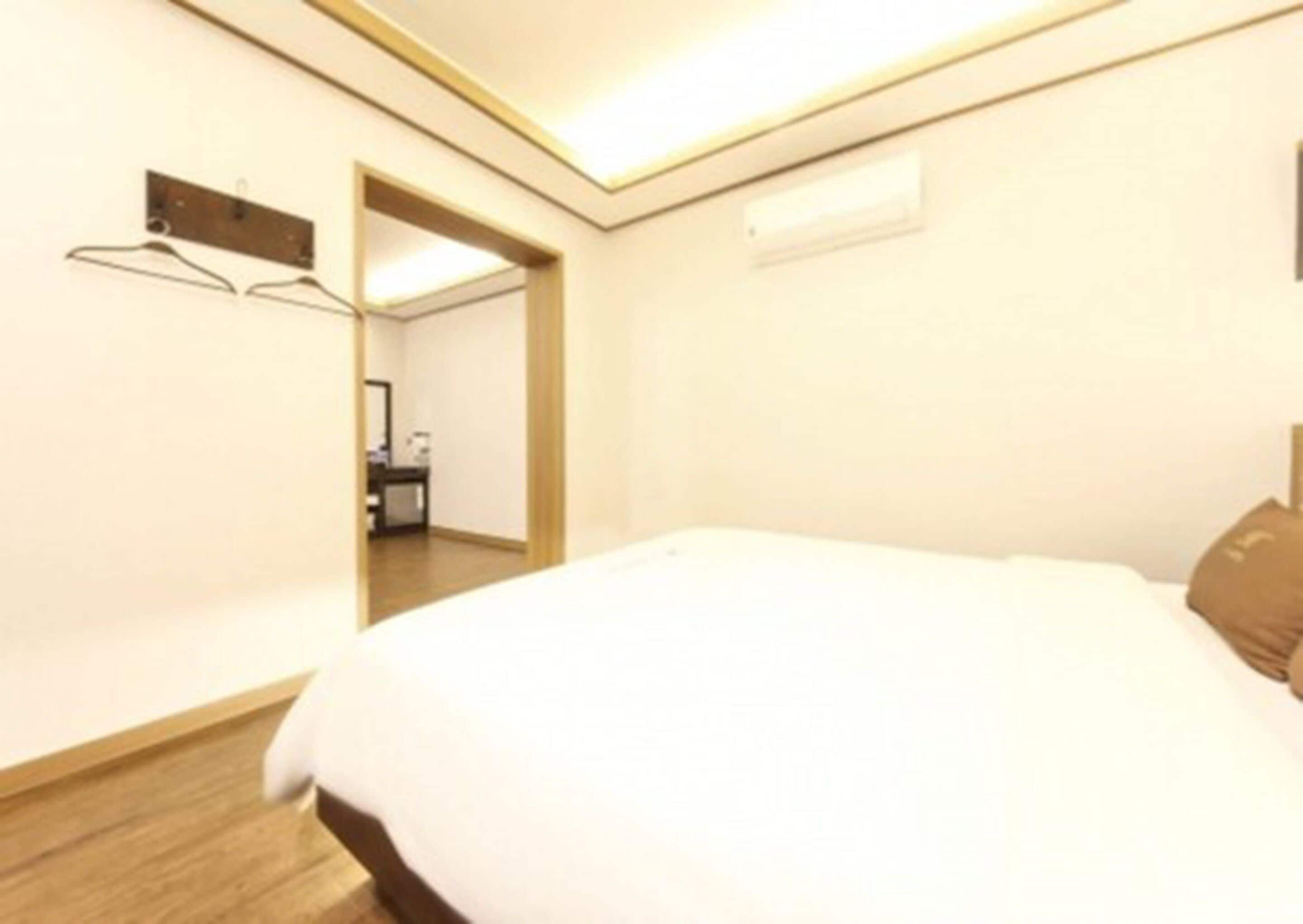 A hotel room with a bed, a wooden floor, a mirror, and a small table with a lamp.